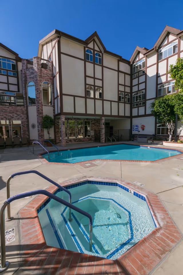 Outdoor courtyard showing a hot tub in the foreground, a swimming pool behind it, and the Tudor-style assisted living building in the background.