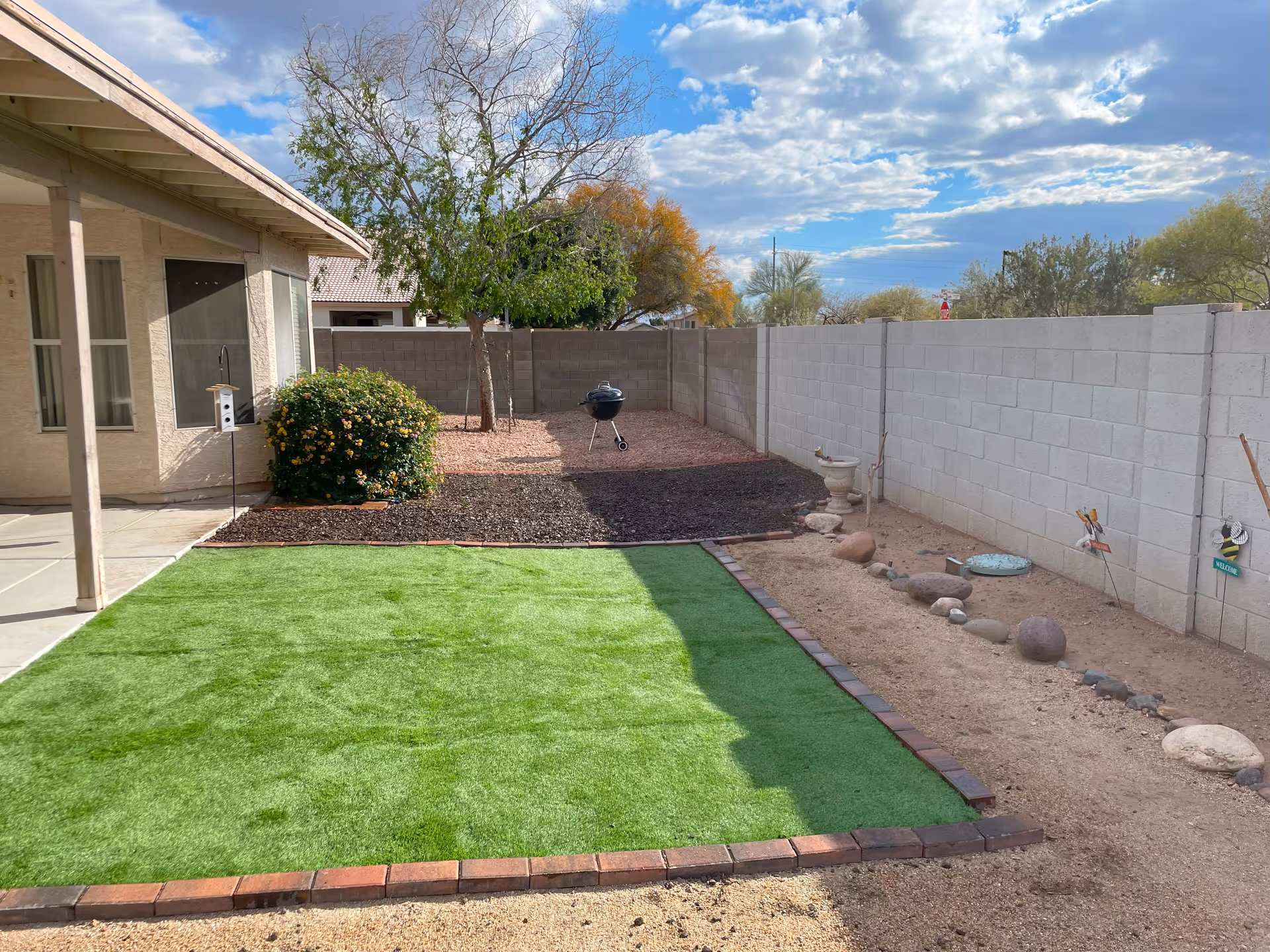 Backyard area of a residential facility with a covered patio on the left, a green artificial grass lawn bordered by bricks, a tree and a bush near a concrete block wall, a charcoal grill, and a clear blue sky with some clouds.
