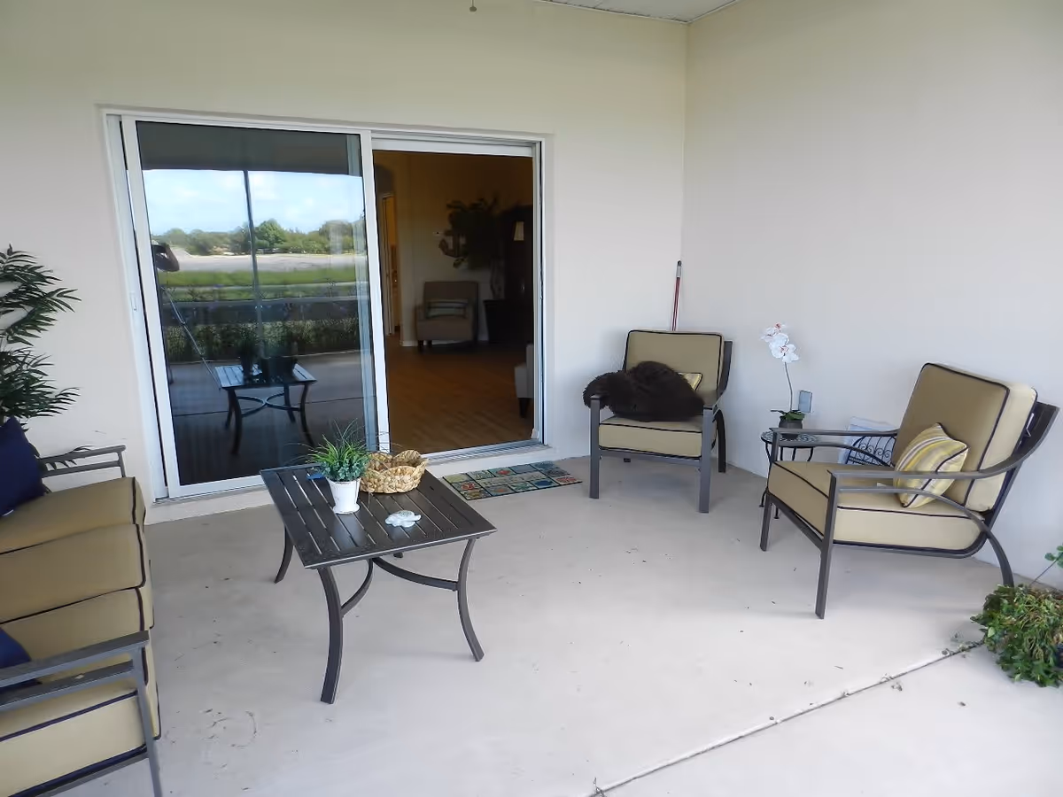A covered outdoor patio area with beige cushioned metal chairs and a matching loveseat arranged around a black metal coffee table. The table has small potted plants and a basket on it. A sliding glass door leads inside to a room with wooden flooring and a chair visible. There are plants on the patio and a broom leaning against the wall.