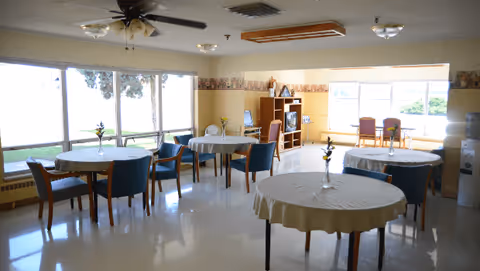 Dining room with round tables covered in white tablecloths, blue chairs, large windows, and a ceiling fan.