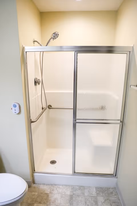 A small shower stall with sliding frosted glass doors and a handheld showerhead mounted on the wall. There is a horizontal grab bar inside the shower. The floor is tiled, and a portion of a toilet is visible on the left side.