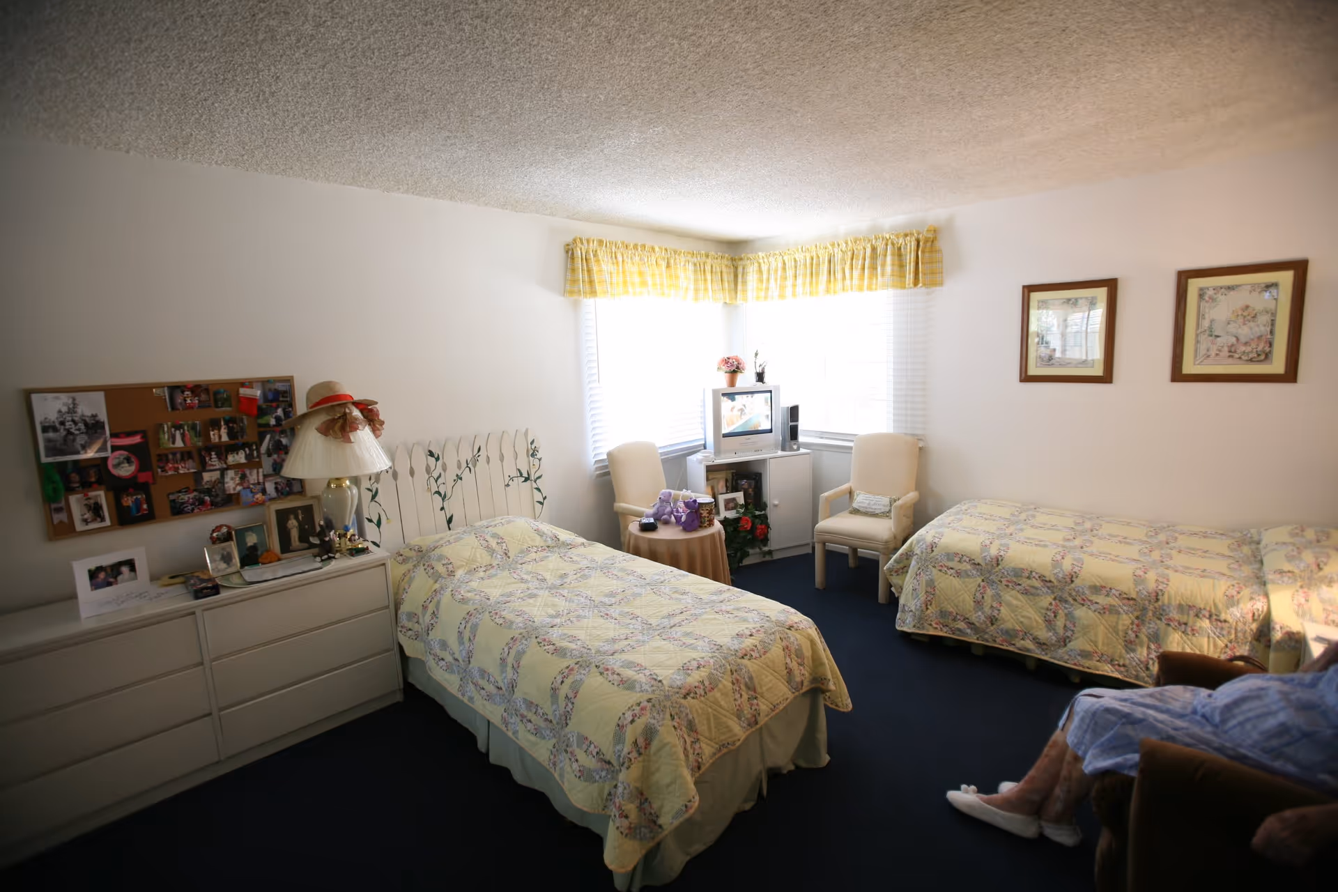 A cozy bedroom with two single beds covered in matching yellow floral quilts. Between the beds is a small round table with a purple stuffed animal and a TV on a white cabinet in the corner. Two beige armchairs are positioned near the window with yellow plaid valances. A dresser with a lamp, photos, and a corkboard with pictures hangs on the wall. An elderly person wearing slippers and a blue robe is seated in a brown armchair on the right side of the room.