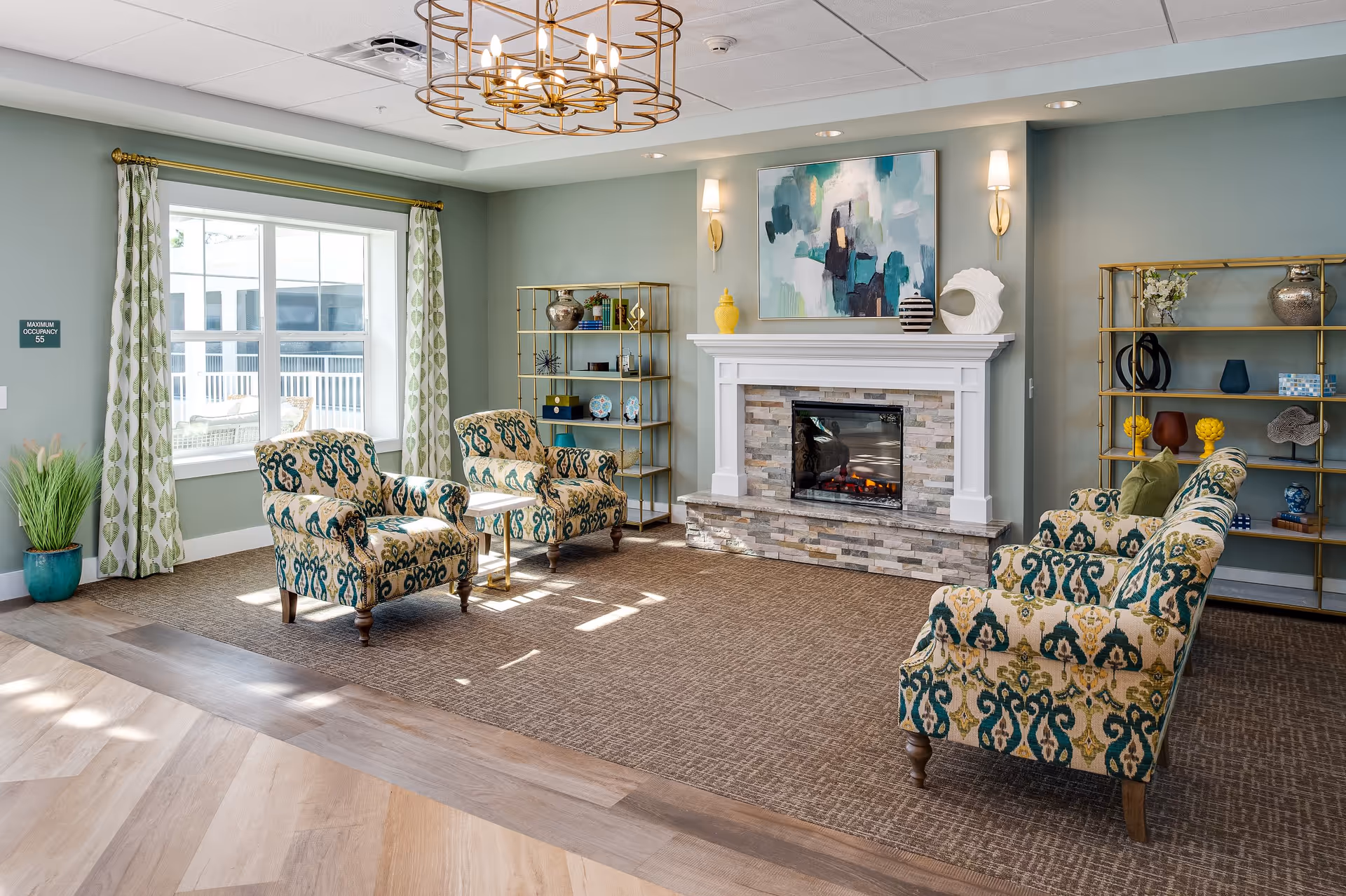 Bright senior living lounge with patterned armchairs arranged around a fireplace, decorative shelving, and a large window.