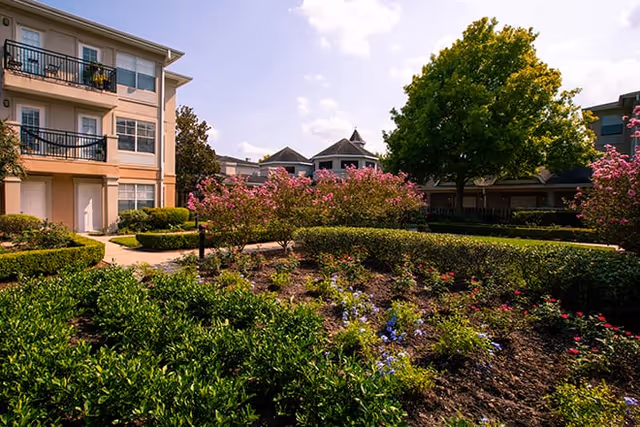 Outdoor garden area at Brookdale Clear Lake with neatly trimmed bushes, flowering plants, and trees. Surrounding buildings with balconies and windows are visible under a partly cloudy sky.