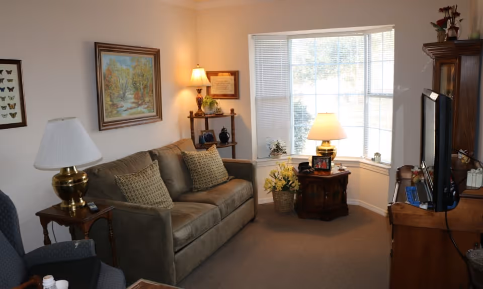 Cozy living room with a sofa, armchair, side tables with lamps, a bay window, and a television.