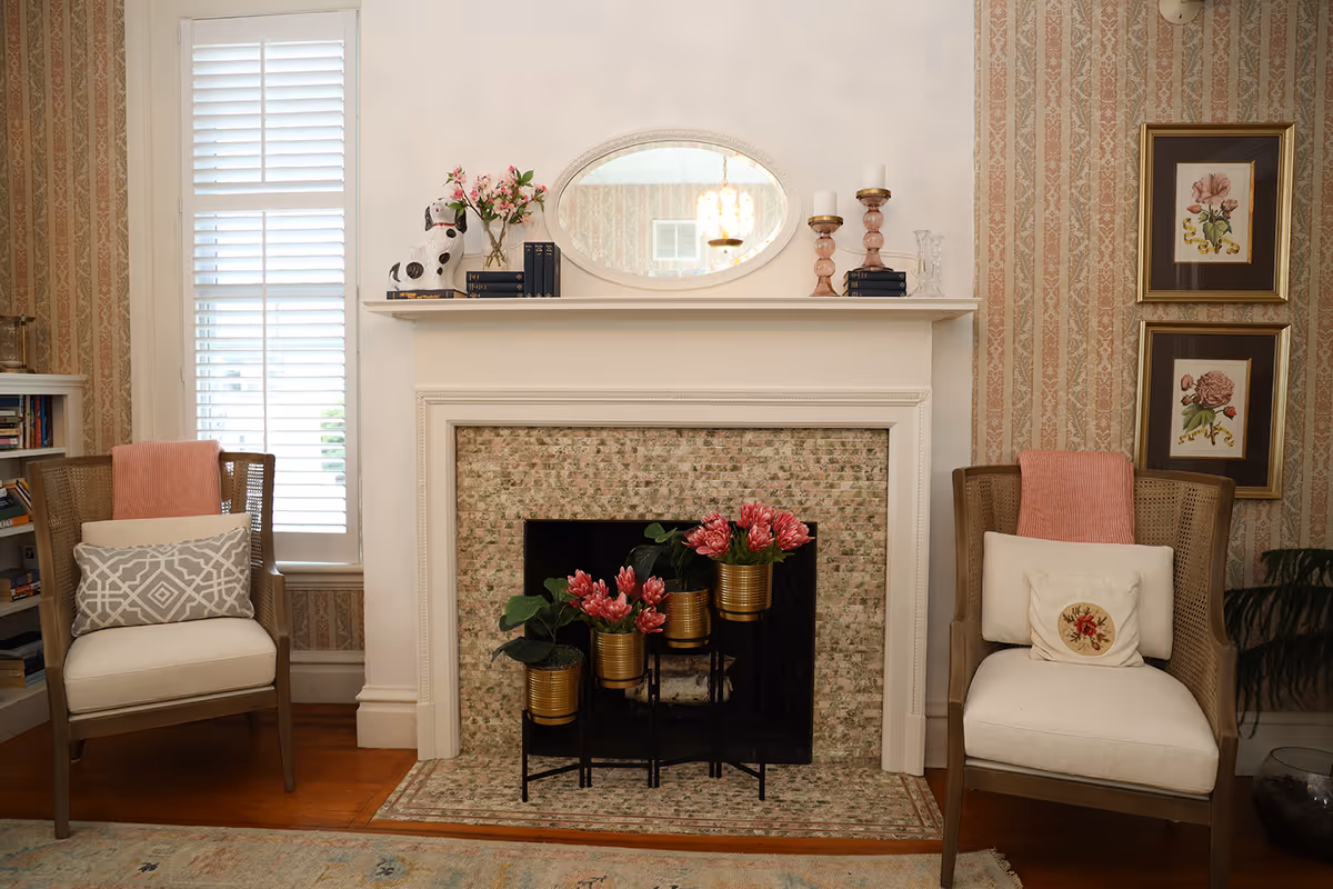 Cozy living room with a decorative fireplace topped by a mirror and candlesticks, flanked by two upholstered armchairs and potted flowers.