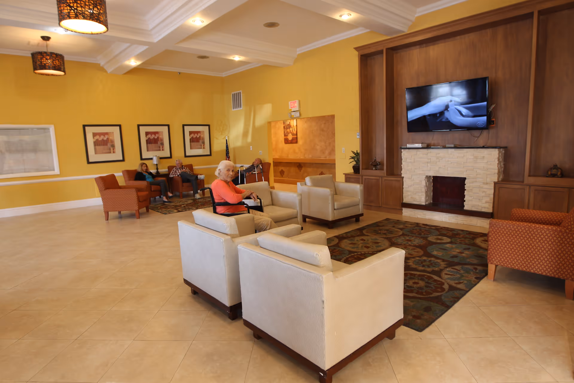 A spacious assisted living facility common area with beige tiled floors, yellow walls, and a white ceiling with recessed lighting. The room features a seating area with cream-colored armchairs arranged around a patterned rug in front of a stone fireplace with a mounted flat-screen TV. In the background, there are additional armchairs and a few elderly residents seated and conversing. Three framed artworks hang on the wall, and an American flag is visible in the corner.