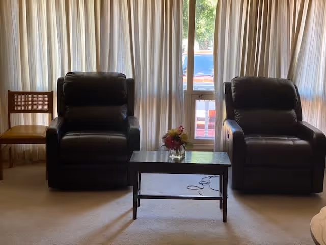 A living room area with two black leather recliner chairs separated by a small dark wooden coffee table with a small flower arrangement on top. To the left of the recliners is a wooden chair with a woven backrest and a yellow cushion. Behind the chairs are large windows covered with light-colored curtains, allowing some natural light to filter through.