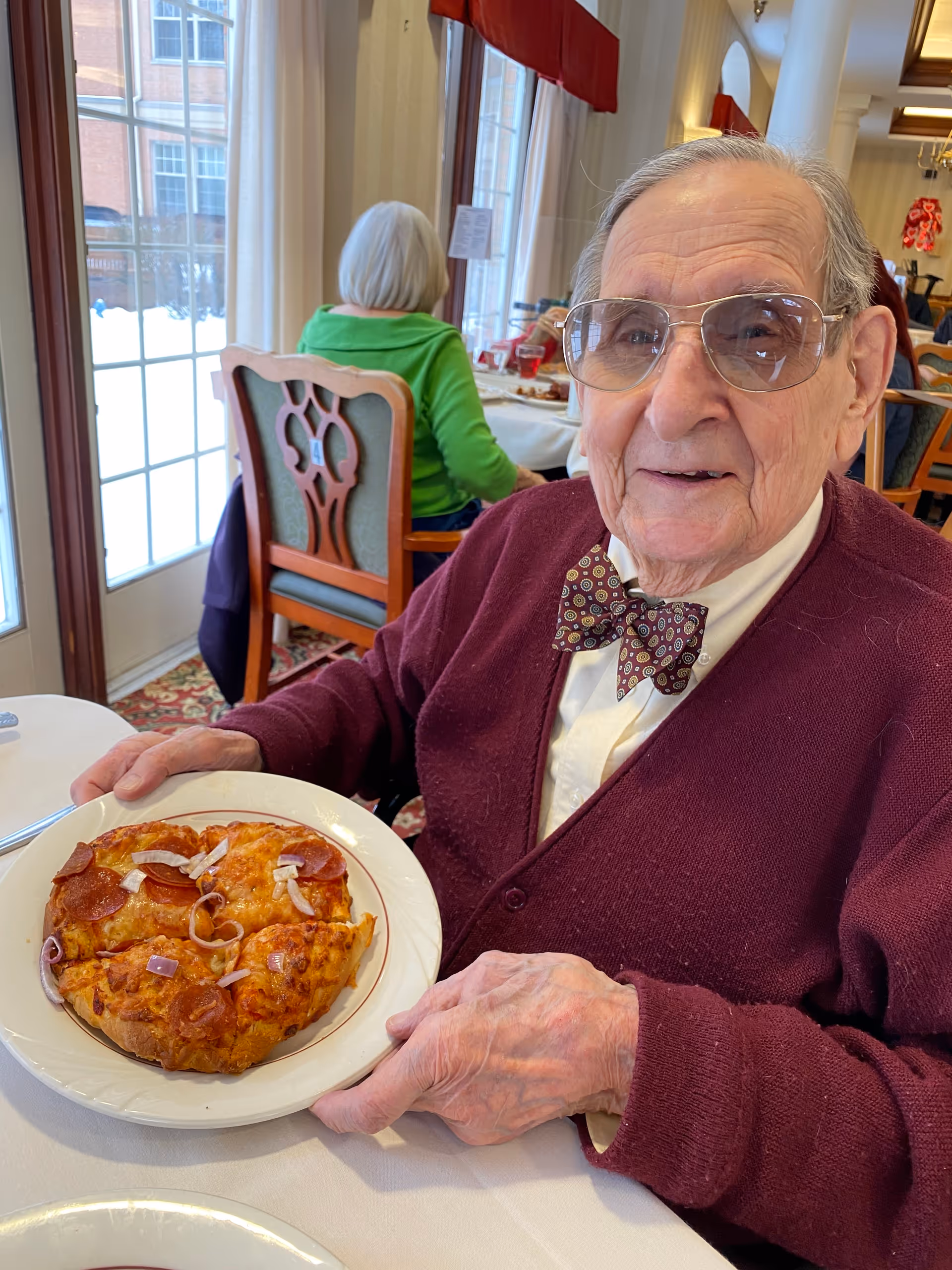 An elderly man wearing glasses, a patterned bow tie, and a maroon cardigan is sitting at a dining table holding a plate with a small pepperoni and onion pizza. Behind him, a woman in a green sweater is seated near large windows with white curtains, and the dining room has wooden chairs and a patterned carpet.