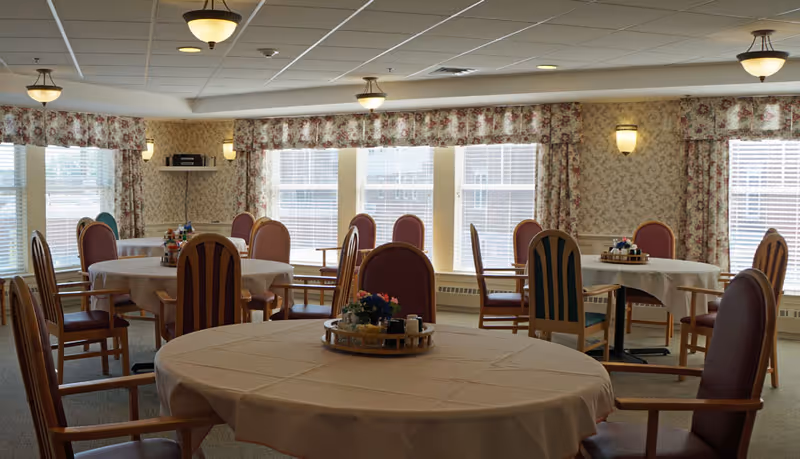 A dining room with multiple round tables covered with white tablecloths, surrounded by wooden chairs with padded seats and backs. The room has large windows with floral curtains, patterned wallpaper, and ceiling light fixtures. Each table has a centerpiece with condiments and flowers.