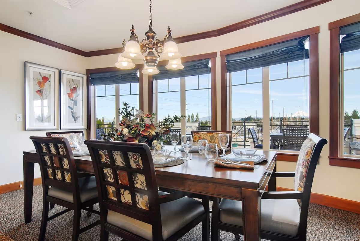 A well-lit dining room with a wooden dining table set for six, featuring patterned cushioned chairs. The table is decorated with a floral centerpiece and glassware. Large windows with dark blinds provide a view of an outdoor patio area and trees beyond. Two framed floral artworks hang on the wall.