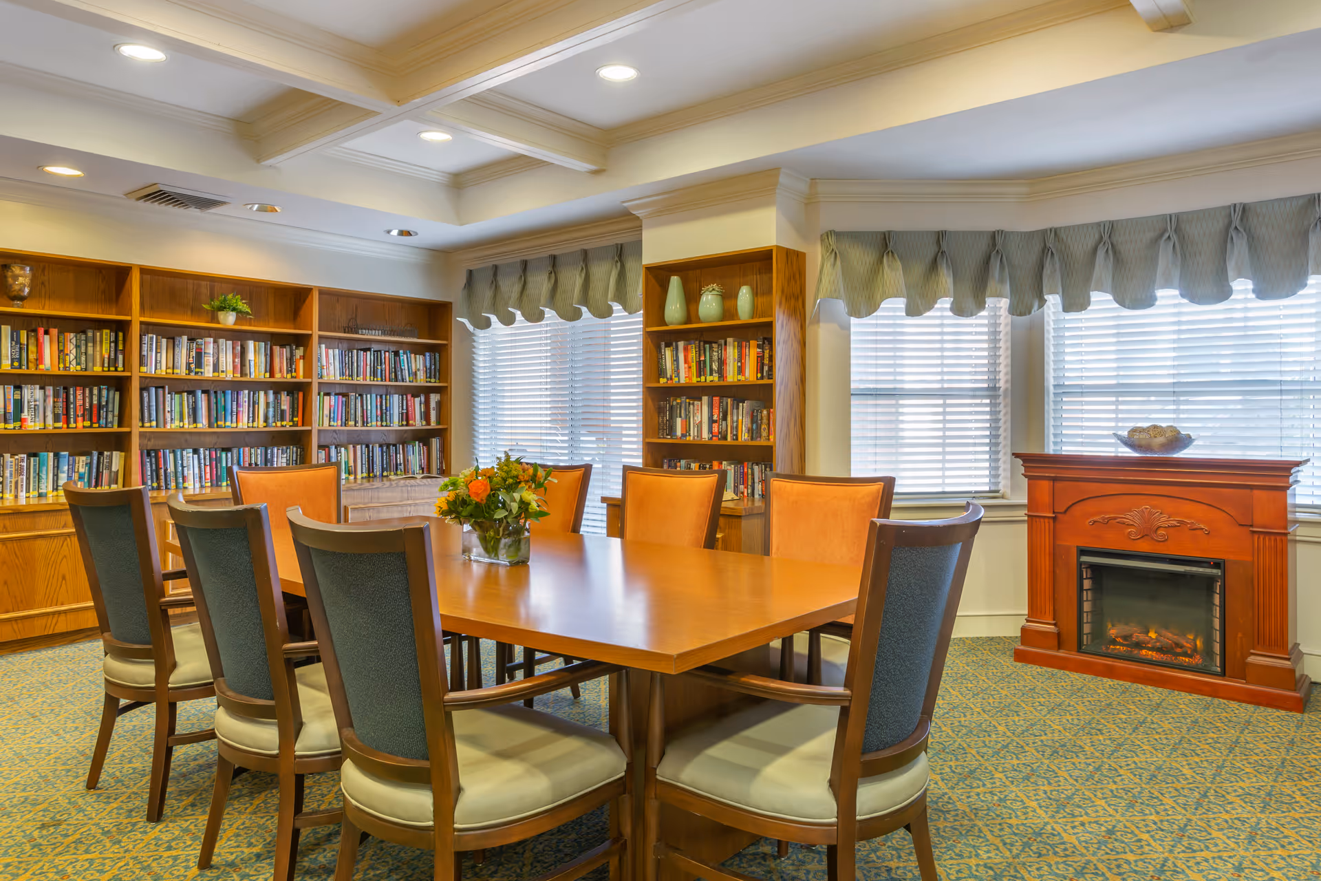 A well-lit communal room with a large wooden table and chairs, bookshelves, windows with valances, and a fireplace.