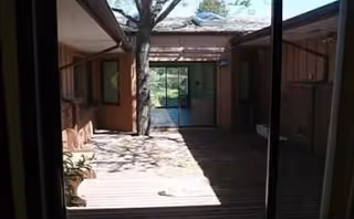 View of an outdoor courtyard area with wooden decking, a tree in the center, and surrounding building walls with windows and doors leading inside.