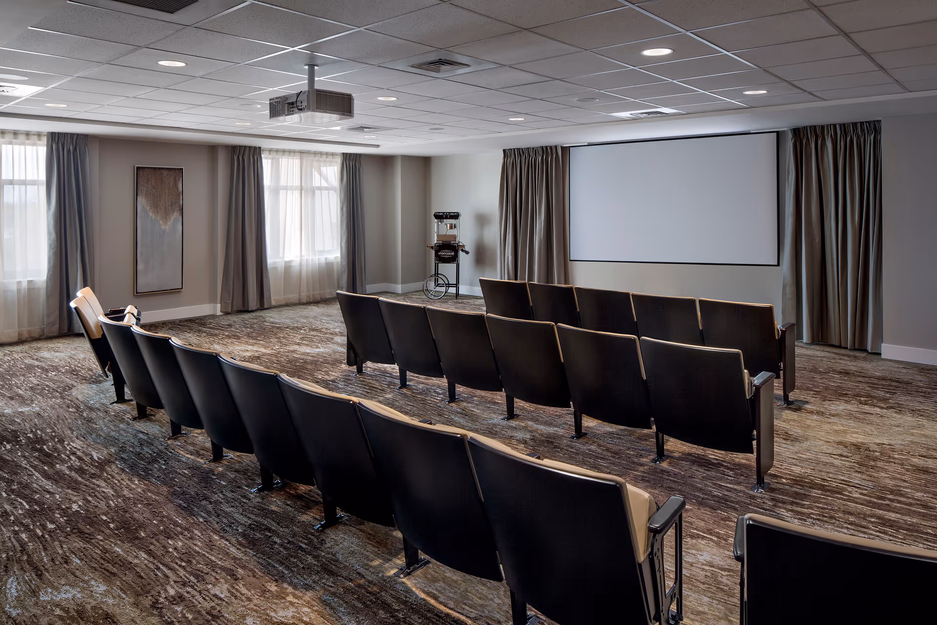 A small theater room with three rows of black cushioned seats facing a large white projection screen. The room has carpeted flooring, two windows with curtains on the left wall, and a popcorn machine in the back corner. A ceiling-mounted projector is visible above the seats.