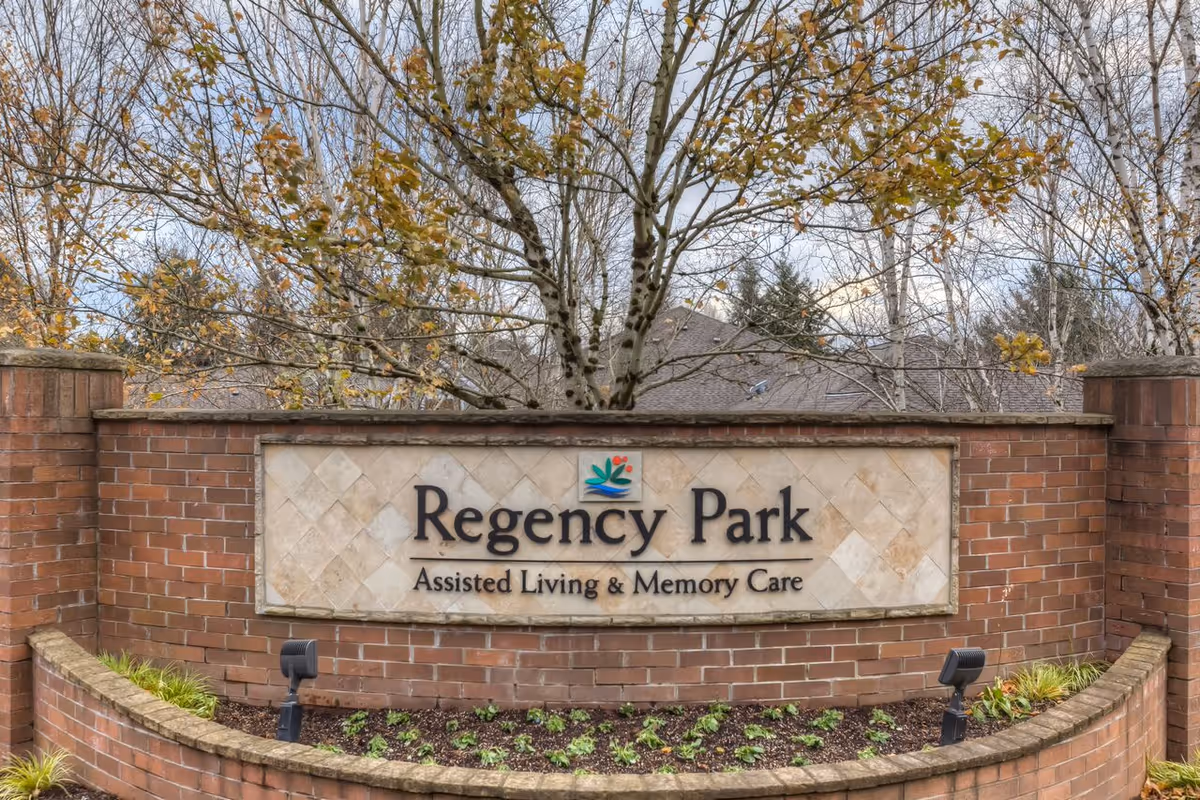 Brick entrance sign for Regency Park Assisted Living & Memory Care with trees and plants in the background.