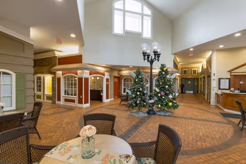 Interior common area of a senior living facility with high ceilings and large windows. The space features brick flooring, several wicker chairs and tables with floral tablecloths, two decorated Christmas trees, and a vintage-style street lamp. The walls have a mix of neutral colors and architectural details resembling storefronts or small building facades.