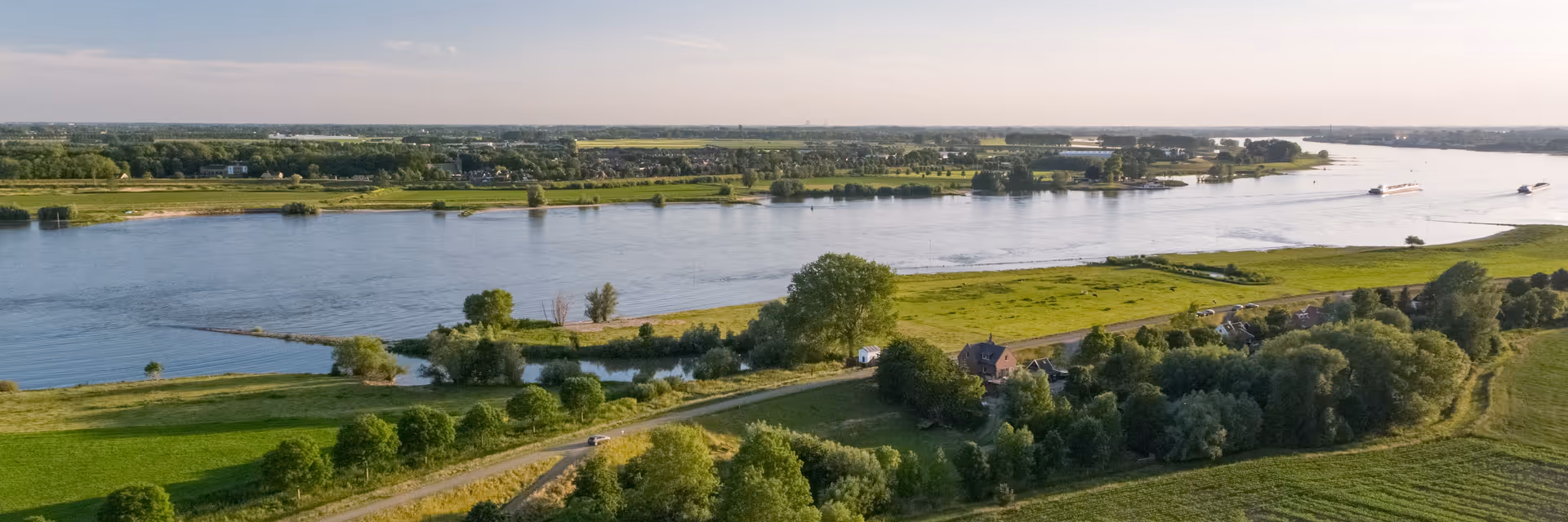 A wide aerial view of a river flowing through a green landscape with trees, fields, and a few houses along the riverbank under a clear sky.