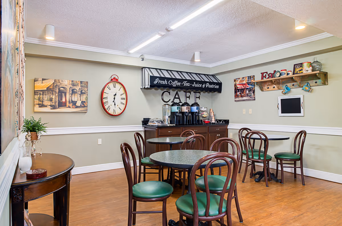 A cozy cafe area inside a senior living facility with round tables and wooden chairs with green cushions. The back wall features a counter with coffee dispensers and a sign above reading 'Fresh Coffee - Tea - Juice & Pastries CAFE'. The walls are decorated with a large red clock, paintings, and a wooden shelf holding mugs and decorative items. The floor is wooden, and the lighting is bright with ceiling fixtures.