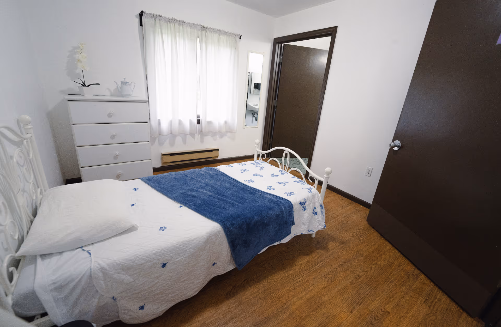 A small bedroom with a single white metal bed frame, a white pillow, and a white quilt with blue floral patterns and a blue blanket folded on top. There is a white chest of drawers with a white teapot and a white orchid plant on it. A window with white curtains is on the wall behind the chest of drawers. The room has wooden flooring and two brown doors, one of which is open, showing a glimpse of a bathroom sink and mirror.