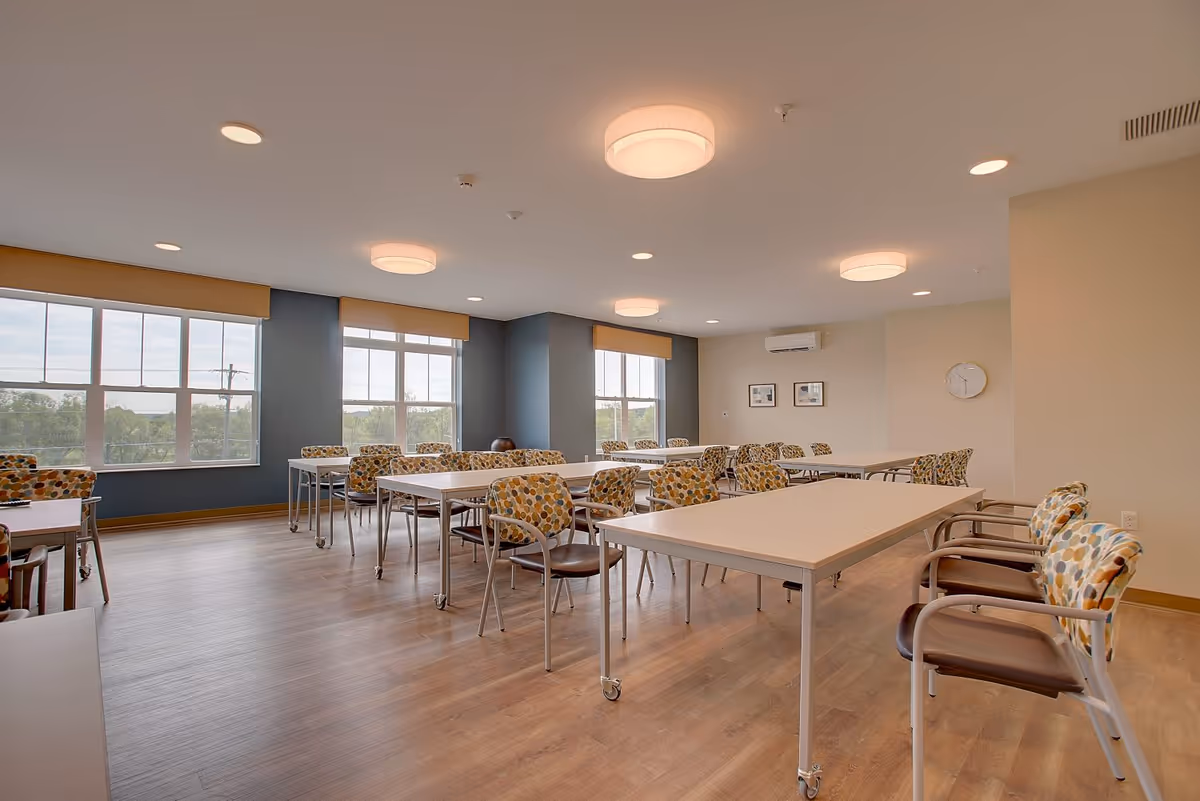 A bright and spacious room with multiple rectangular tables and chairs with colorful patterned upholstery arranged neatly. The room has large windows letting in natural light, light wood flooring, and modern ceiling lights. There is a wall clock and framed pictures on the far wall, and an air conditioning unit mounted above.
