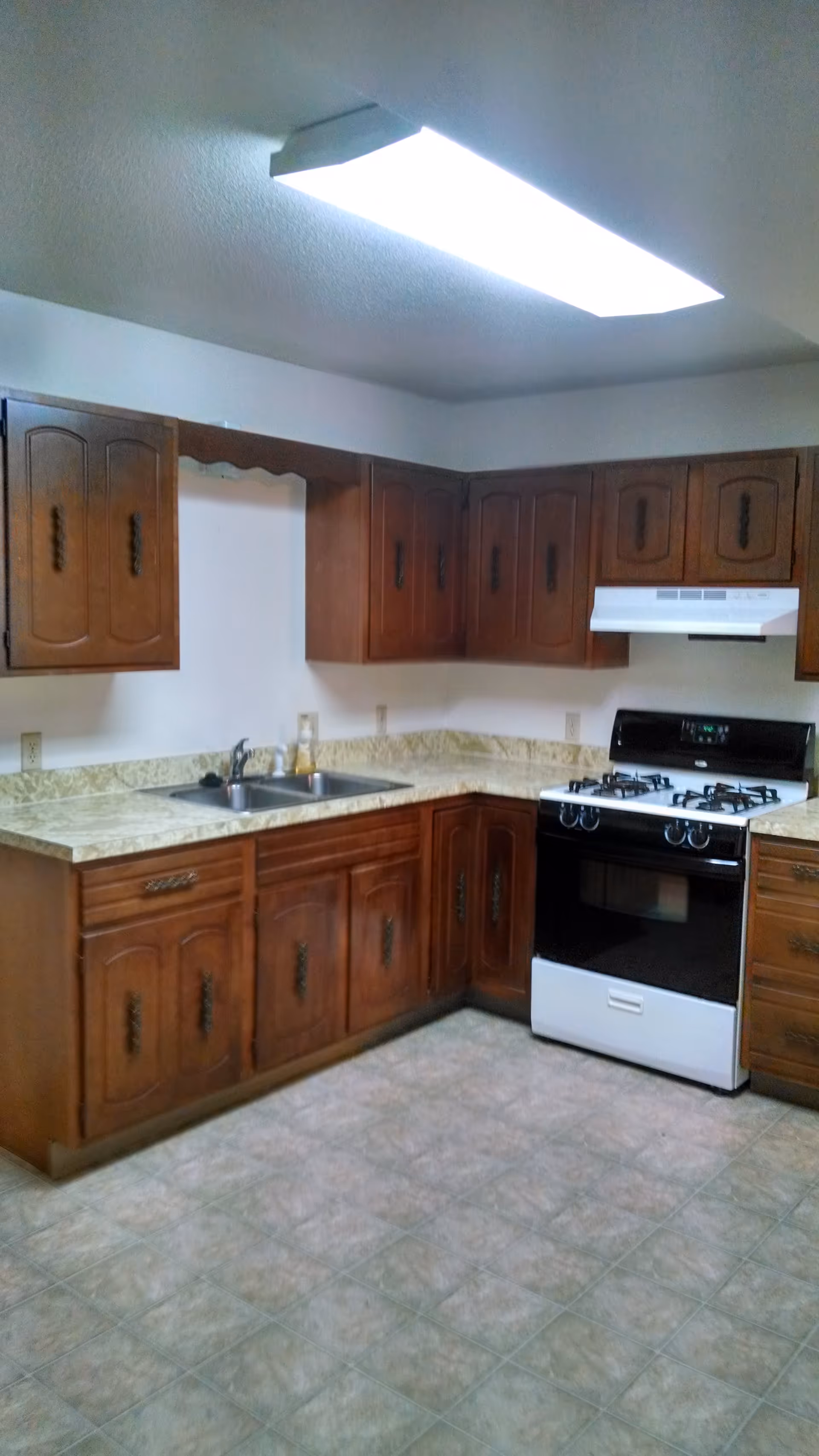 Interior view of a kitchen with wooden cabinets, a double sink, a gas stove with an oven, and a tiled floor. The kitchen has a bright rectangular ceiling light.