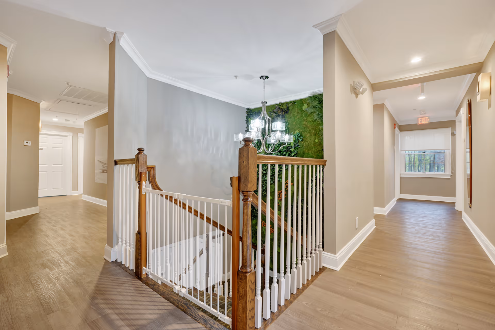 Interior hallway of a senior living facility with light wood flooring, beige walls, white trim, and a staircase with wooden handrails and white balusters. A modern chandelier hangs above the staircase, and a green plant wall is visible in the stairwell. The hallway has a window at the end with a white blind and an exit sign above the door.
