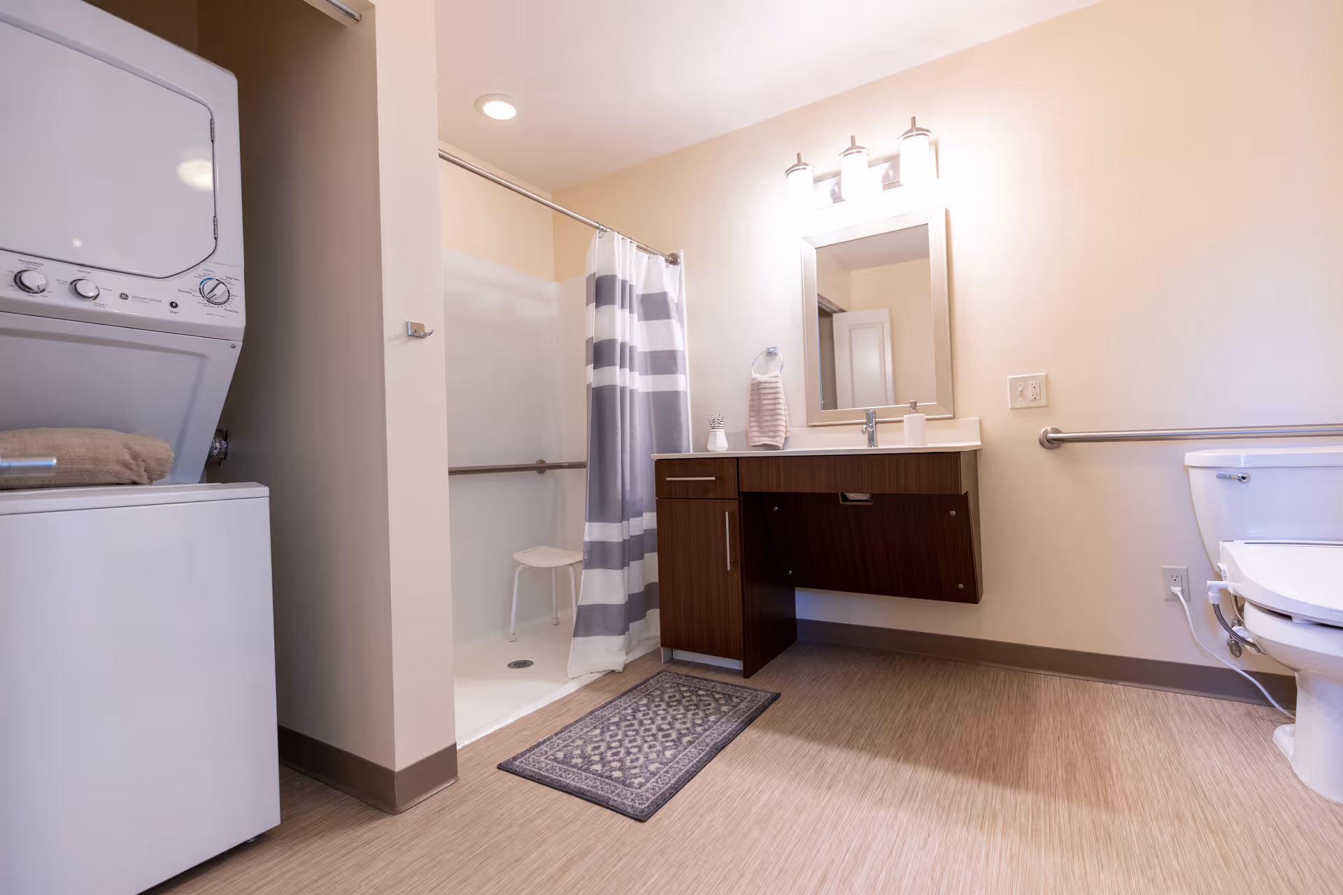 A clean and accessible bathroom featuring a walk-in shower with a striped curtain and a white stool inside. There is a wooden vanity with a sink, a mirror above it, and a towel hanging on a ring. A toilet with a grab bar is visible on the right side. On the left side, there is a stacked washer and dryer unit with a folded towel on top. The floor has a patterned rug in front of the vanity.