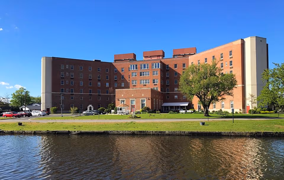 Large brick senior living building sits along a river with a green lawn and trees.