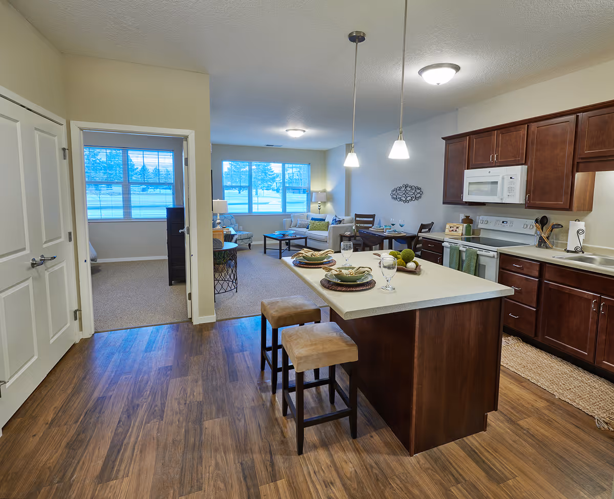 Open-plan kitchen with a center island and two stools opening into a bright living room with large windows.