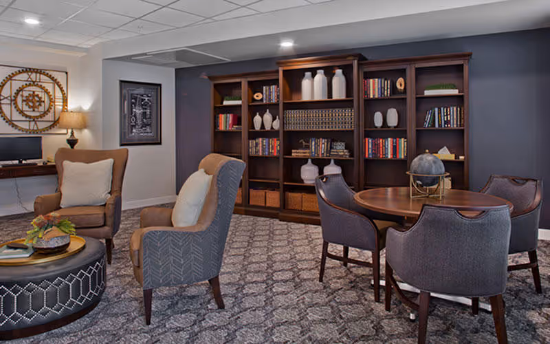 A cozy living room area with patterned carpet, two upholstered armchairs with cushions, a round wooden table surrounded by four gray chairs, and a large wooden bookshelf filled with books and decorative items against a dark blue wall. A lamp and wall art are visible in the background.