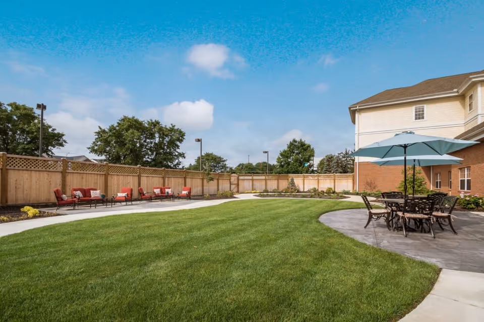 Outdoor patio area at The Forum At Knightsbridge featuring a well-maintained green lawn, a curved concrete pathway, several red cushioned chairs lined up along a wooden fence, and a round table with chairs under a large blue umbrella next to a brick building.