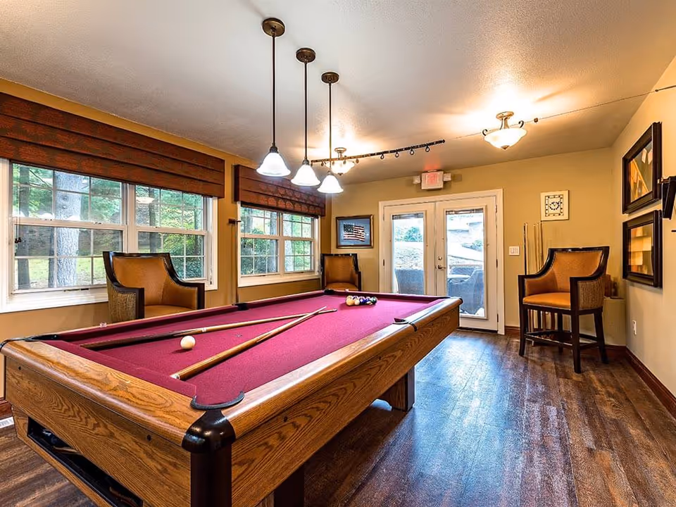 A well-lit game room with a pool table featuring a red felt surface. Two pool cues and a white cue ball are on the table, with a rack of billiard balls at one end. The room has wooden flooring, beige walls, three pendant lights hanging above the pool table, and two large windows with brown blinds. There are three cushioned chairs placed around the room, framed artwork on the walls, and a set of double glass doors leading outside.