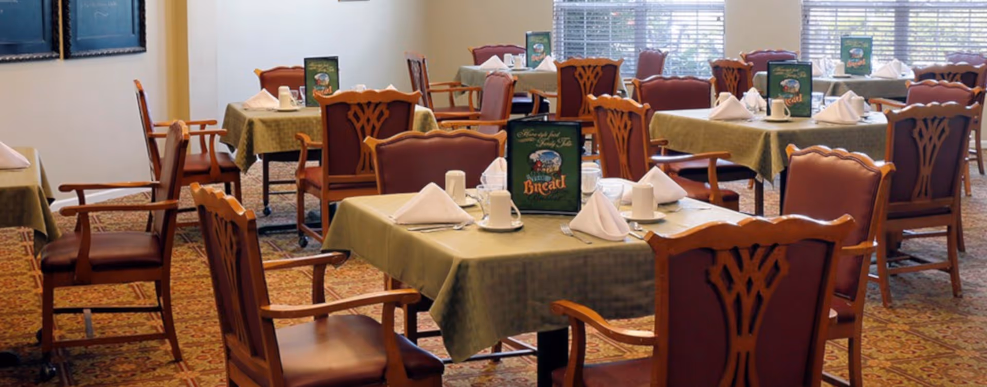 A dining room with several tables covered in green tablecloths, each set with white napkins, cups, glasses, and menus. The chairs are wooden with cushioned seats and backs. Large windows with blinds allow natural light to fill the room.