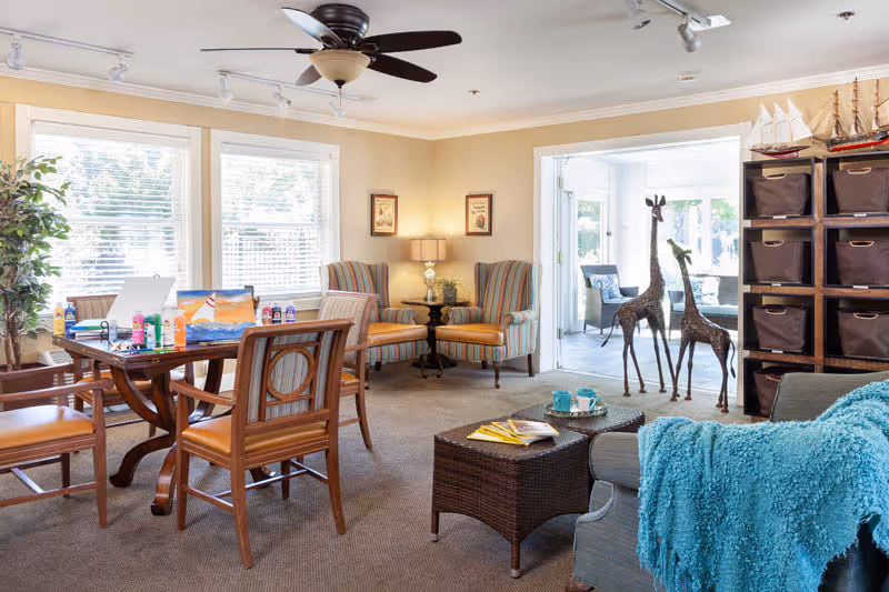 A bright and cozy living room area with a ceiling fan, large windows with blinds, a wooden table with chairs and art supplies, two striped armchairs with a small table and lamp between them, a wicker coffee table with magazines and cups, a gray sofa with a blue throw blanket, a shelving unit with brown storage bins, and two decorative giraffe sculptures near a sunroom with glass doors.