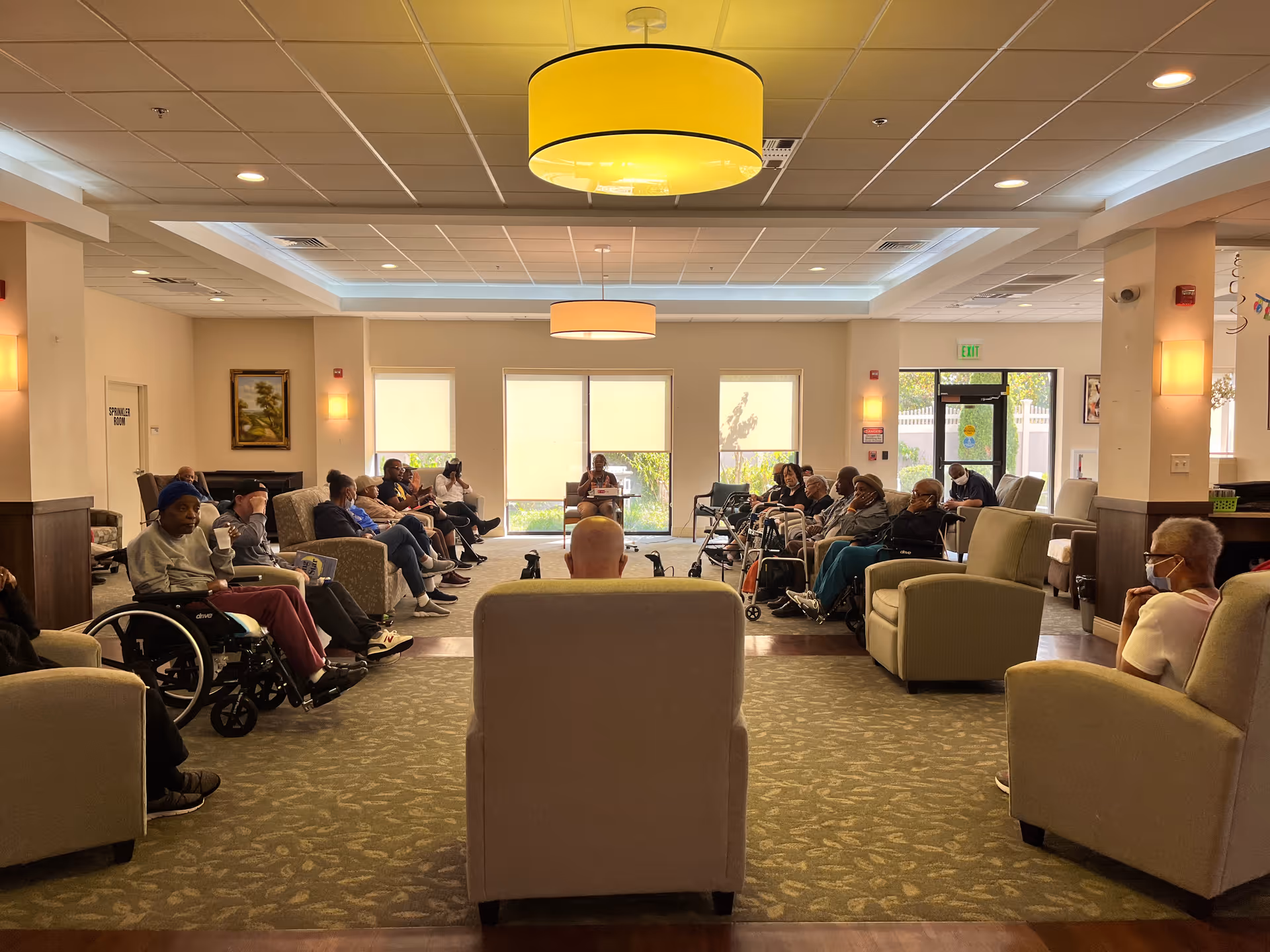 A large room with many elderly individuals seated in a semi-circle on chairs and wheelchairs, facing a person at the front of the room. The room has beige walls, large windows with blinds, and modern ceiling lights. Some people are wearing masks.