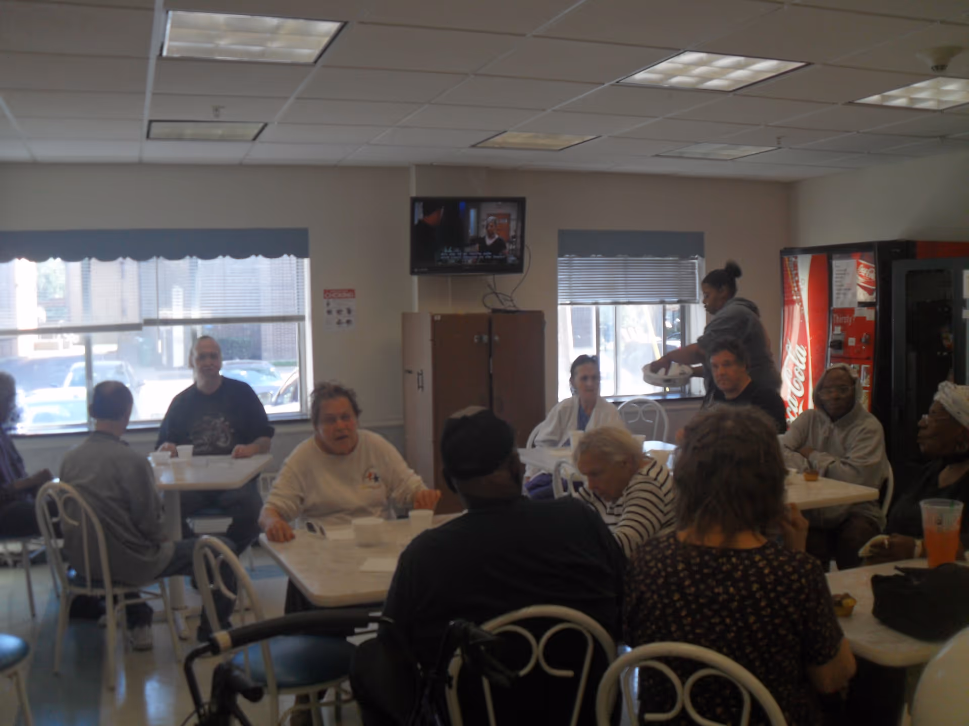 A group of elderly people sitting around tables in a communal dining area with white chairs and tables. A woman is serving food near a window, and a television is mounted on the wall above a cabinet. There is a Coca-Cola vending machine in the corner of the room.