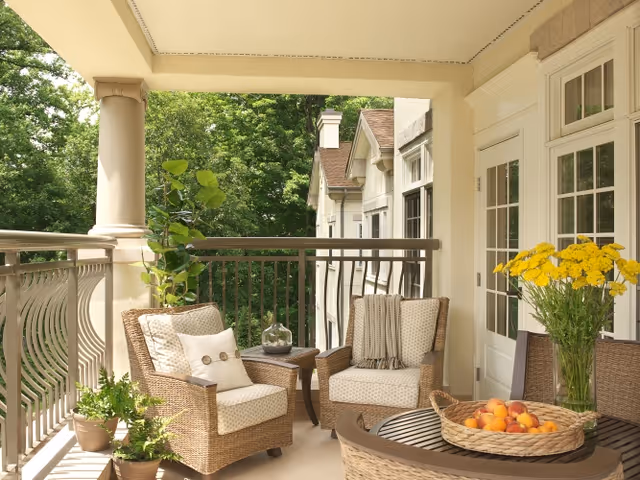 A cozy outdoor balcony with two wicker armchairs featuring cushions and a throw blanket, a small side table with a decorative item, a round table with a basket of peaches, a vase of yellow flowers, and several potted plants. The balcony overlooks lush green trees and is attached to a building with cream-colored walls and large windows.