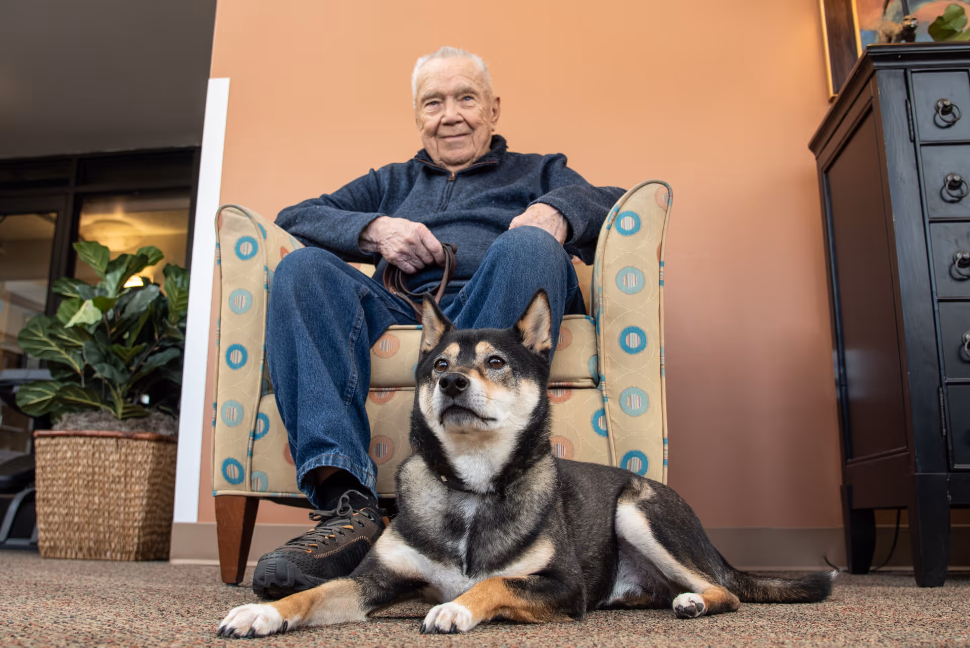 An elderly man sitting in a patterned armchair indoors with a black and tan dog lying on the carpet in front of him. There is a plant in a woven basket to the left and a dark wooden cabinet to the right against a peach-colored wall.