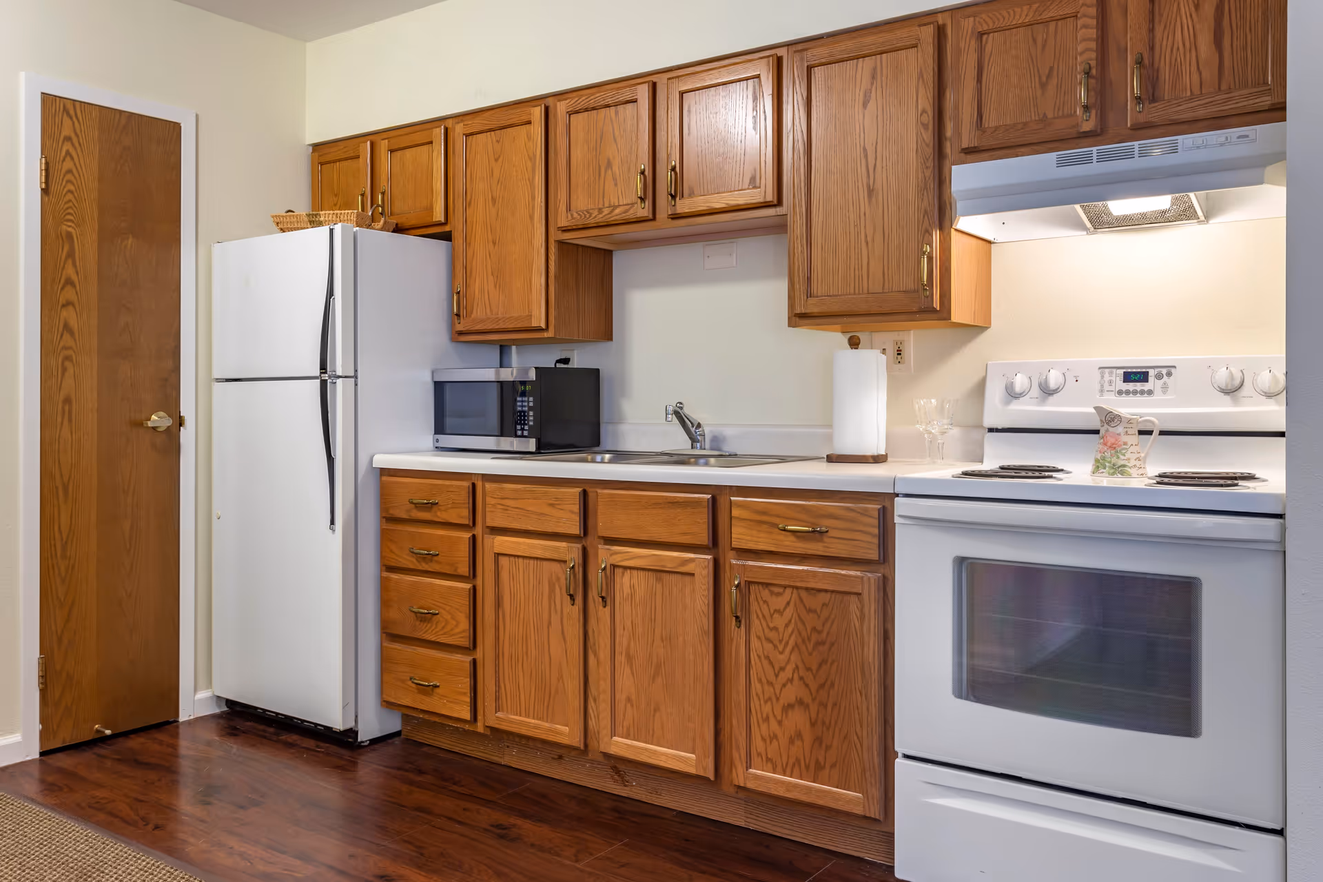 A kitchen with wooden cabinets, a white refrigerator, a microwave on the counter, a double sink, a white electric stove with a floral pitcher on top, and a paper towel holder. The floor is dark wood and there is a wooden door to the left.