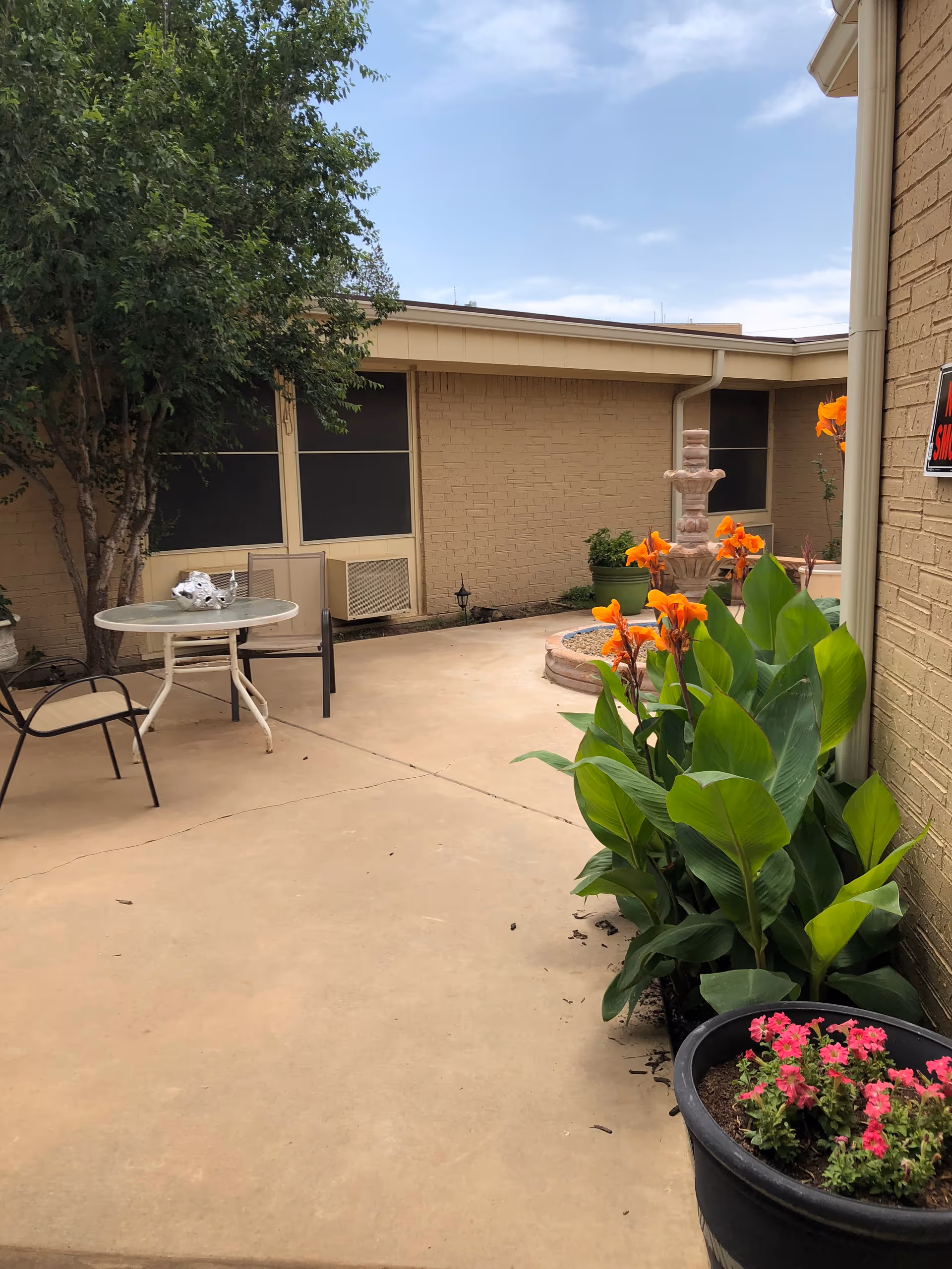 Outdoor courtyard area with a concrete floor, a round glass table with two chairs, potted plants with orange and pink flowers, a tree, and a stone water fountain against a beige brick building under a partly cloudy sky.