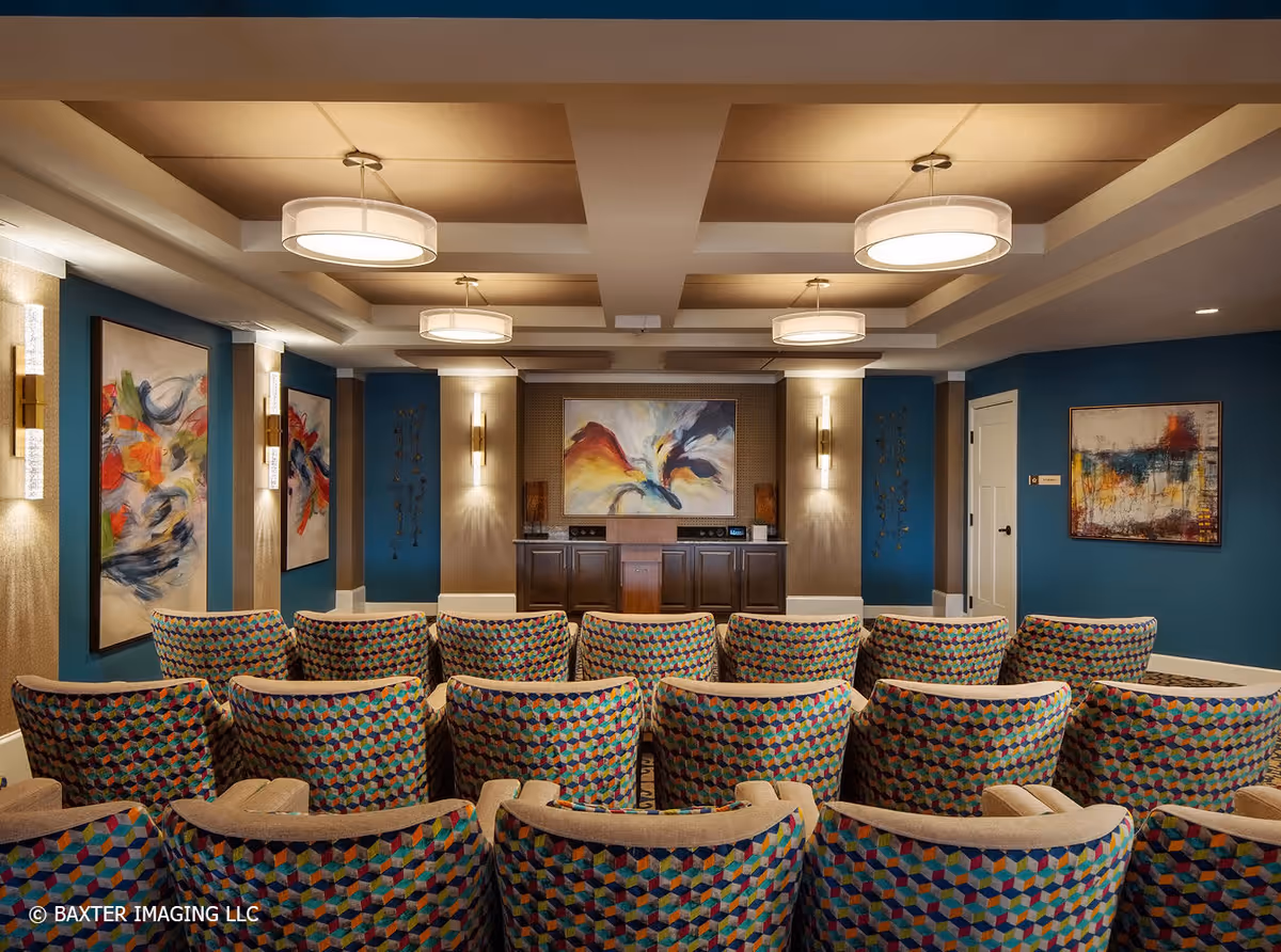 A cozy theater-style room with multiple rows of colorful patterned chairs facing a wall with abstract paintings and a wooden cabinet. The room has blue walls, modern ceiling lights, and wall sconces providing warm lighting.