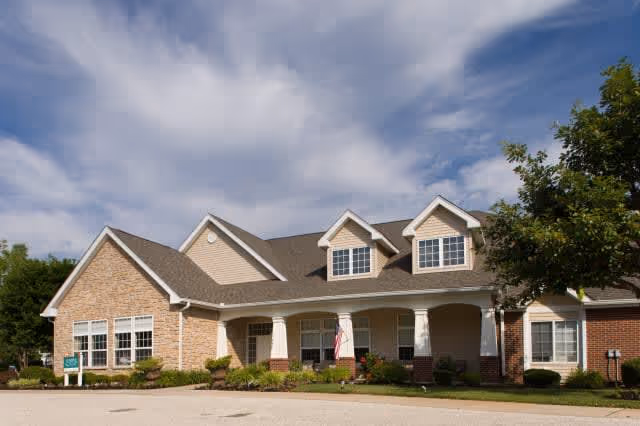 Exterior view of a single-story senior living facility building with a combination of brick and siding, featuring a covered entrance with columns, multiple windows, and a well-maintained landscape under a partly cloudy sky.