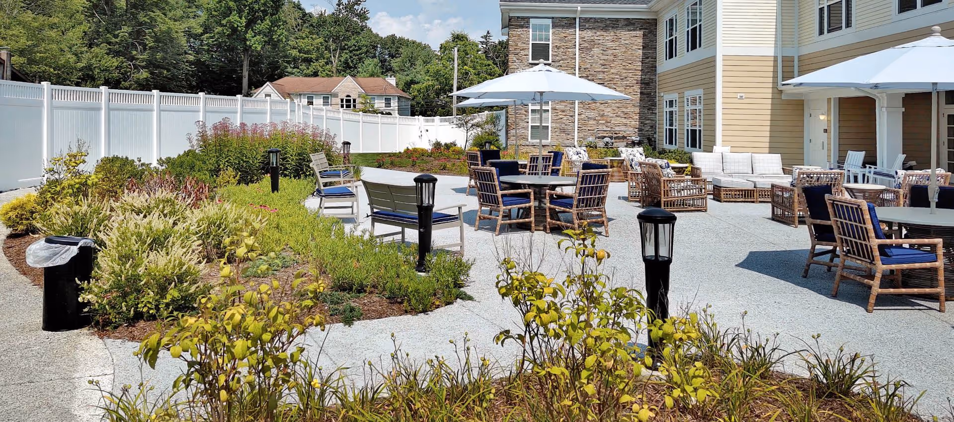 Outdoor courtyard patio with tables, umbrellas, cushioned chairs and landscaped plant beds beside a senior living building.
