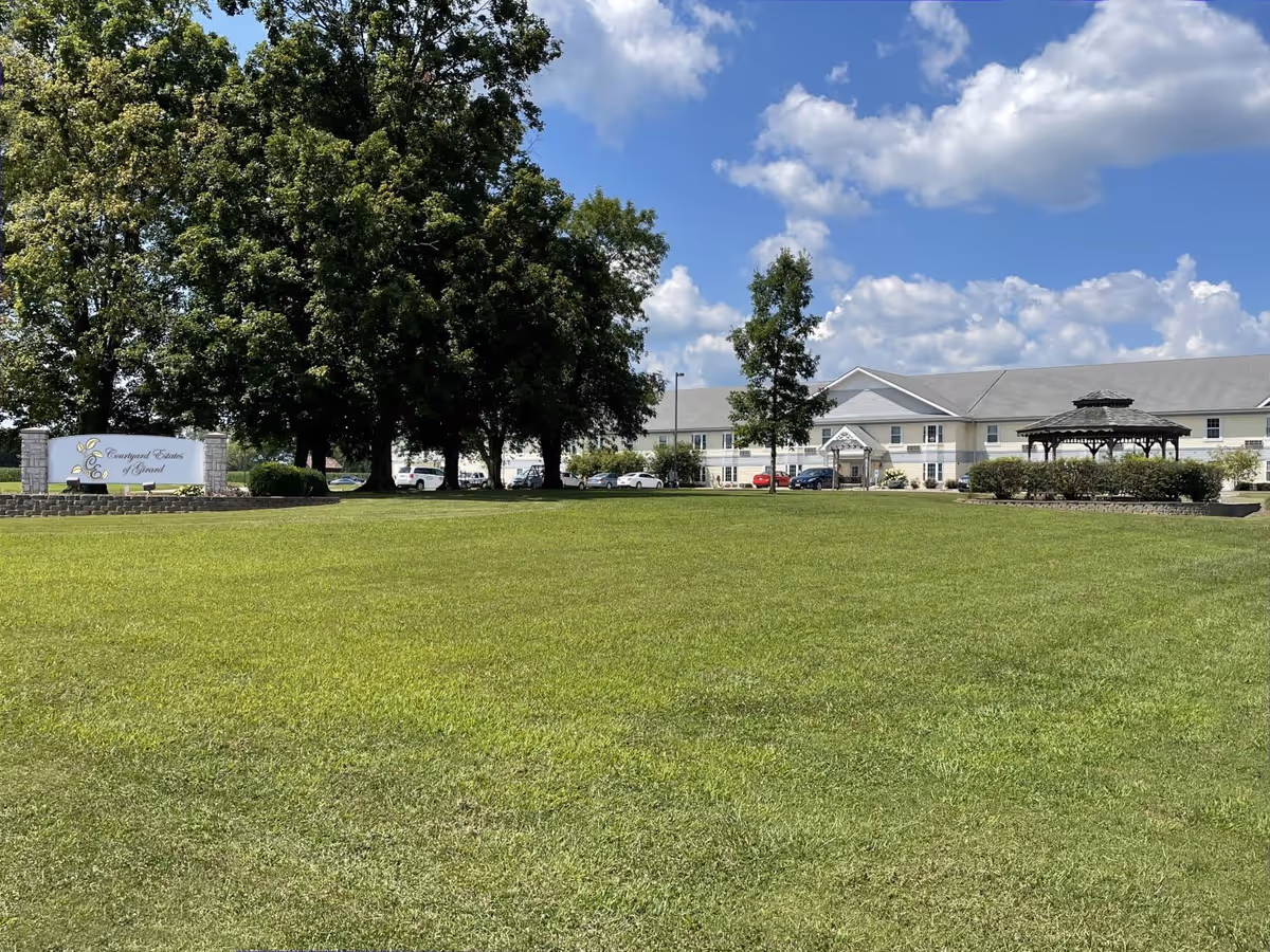 Wide view of Courtyard Estates of Girard facility showing a large green lawn in the foreground, a few tall trees, a white building with multiple windows in the background, a gazebo on the right side, and a sign with the facility name on the left side under a partly cloudy blue sky.