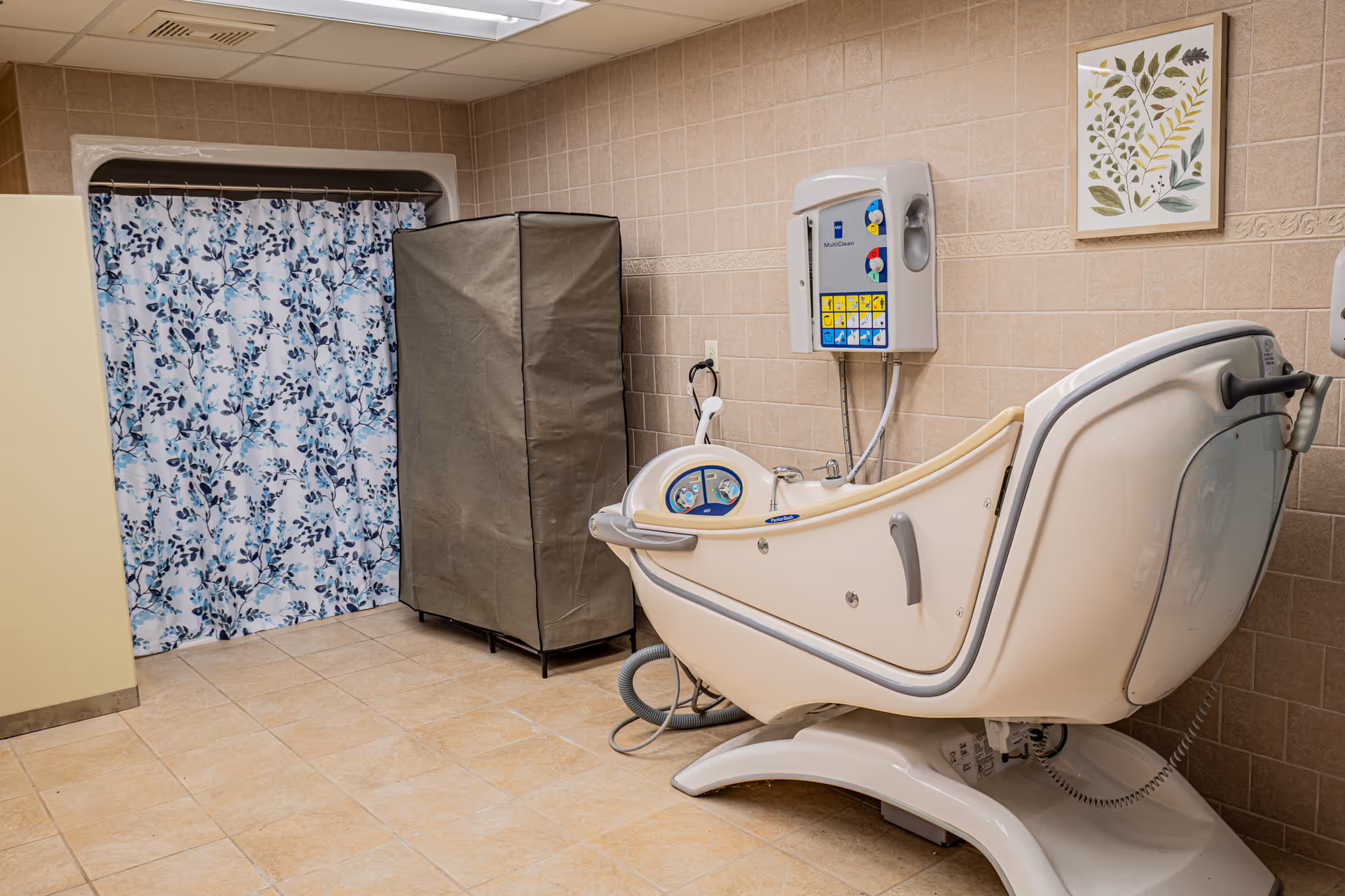 A tiled room with a specialized bathing chair designed for assisted bathing, a floral patterned shower curtain, a covered storage unit, and a wall-mounted control panel. A framed botanical print hangs on the wall.