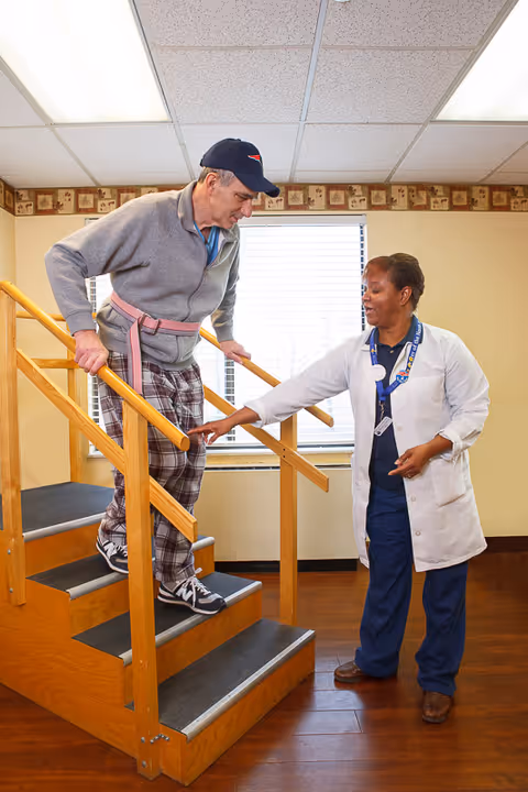 An elderly man wearing a gray sweater, plaid pants, and a cap is walking down a small wooden staircase with handrails on both sides, assisted by a female healthcare worker in a white coat and navy scrubs inside a room with wooden flooring and a window with blinds.