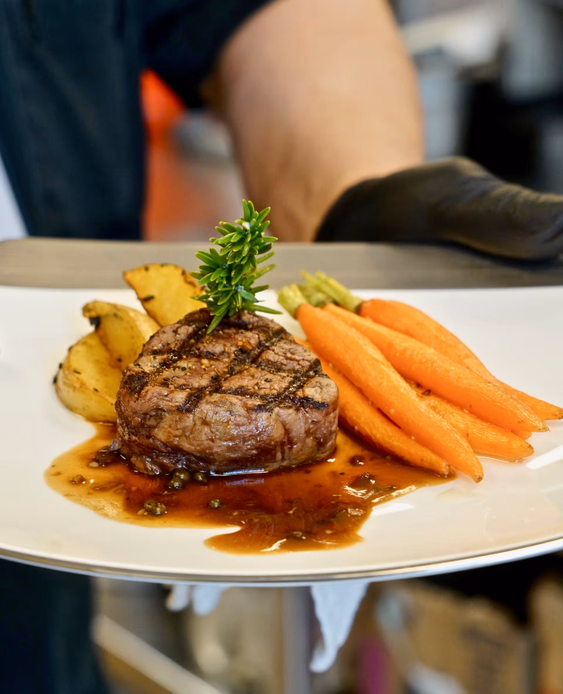 A plated meal featuring a grilled steak garnished with a sprig of rosemary, served with roasted potato wedges and steamed baby carrots, presented on a white plate.