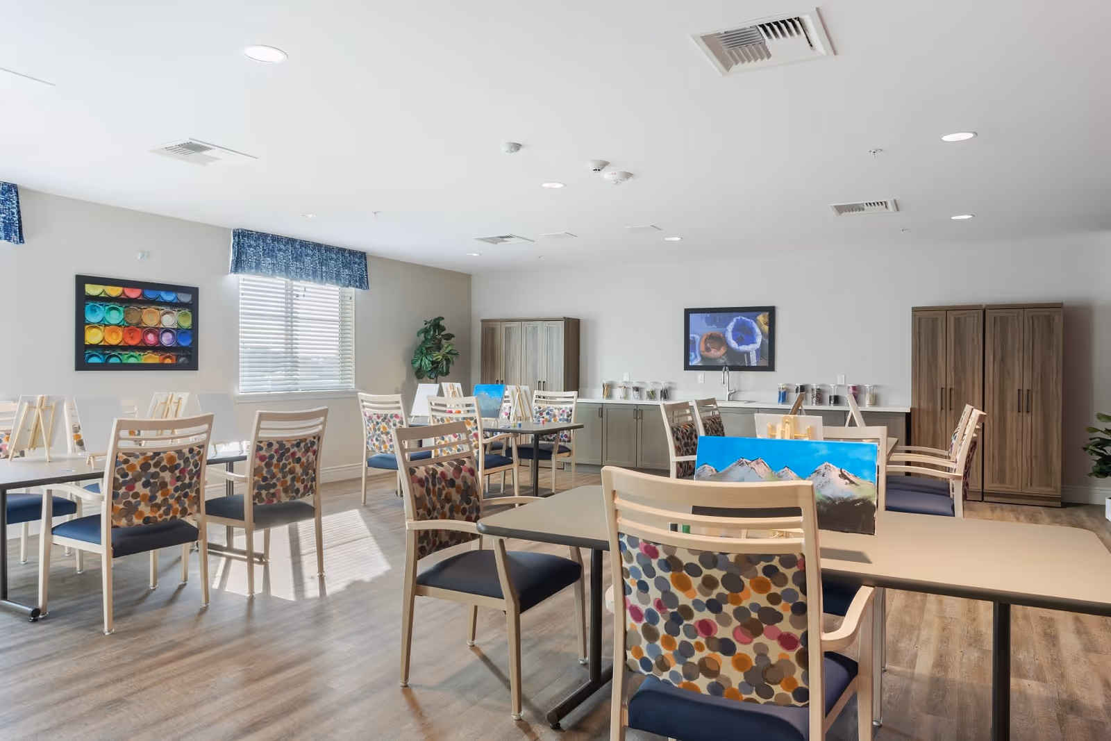 Bright communal activity/dining room with multiple tables and patterned chairs set up for painting activities.