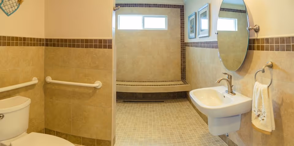 A clean and accessible bathroom with beige tiled walls and floor. The bathroom features a white toilet with grab bars on the walls, a wall-mounted sink with a faucet, a round mirror above the sink, a towel ring with a white towel, and a walk-in shower area with a built-in bench and a window above it.
