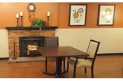 A cozy interior room with a brick fireplace, a wooden mantel decorated with candles and a clock, two framed floral pictures on the wall, and a wooden table with two chairs on a hardwood floor.
